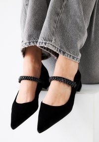 Zapatos Mary Jane de ante negro con punteras afiladas y un tacón pequeño, que presentan una correa decorativa de cuentas negras a lo largo del empeine.