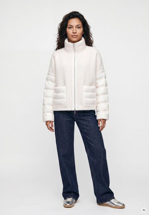 Frau steht und trägt eine weiße Steppjacke, dunkelblaue Jeans mit weitem Bein und silberne Sneakers vor einem schlichten hellen Hintergrund.