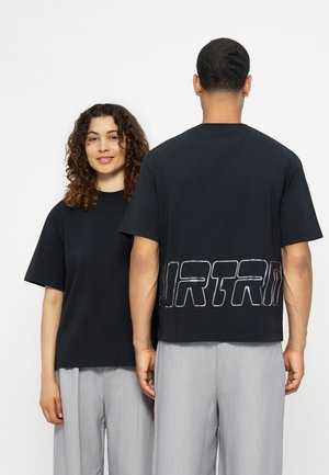 Δύο άτομα φορούν μαύρα φαρδιά t-shirts και ανοιχτό γκρι παντελόνια· το ένα κοιτάει προς τα εμπρός και χαμογελά, ενώ το άλλο δείχνει μια εκτύπωση στην πλάτη με μεγάλα γράμματα.