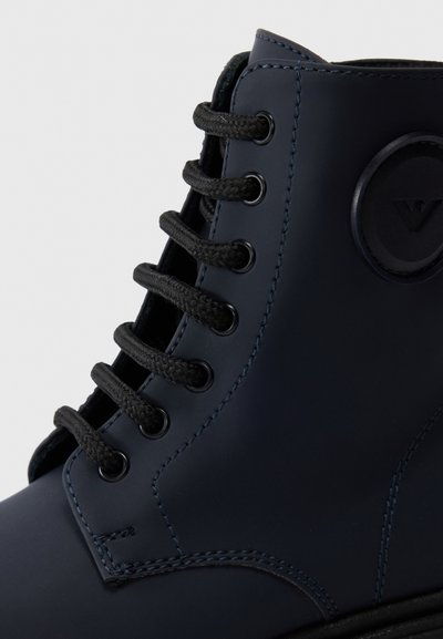 Stivaletto blu navy con materiale sintetico liscio, lacci neri, occhielli rotondi e un'accattivante etichetta circolare sul lato. Suola in gomma spessa.