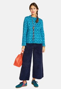 Türkise gemusterte Bluse mit langen Ärmeln, marineblaue weite Cord-Hose, orangefarbige Handtasche, türkisfarbene und rosafarbene Sneakers sowie eine Perlenkette.