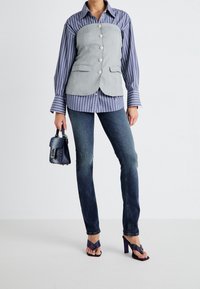 Chemise rayée bleue superposée sous un gilet gris à boutons, associée à un jean en denim noir et des sandales à talons violettes. Petit sac à main noir avec des accents.