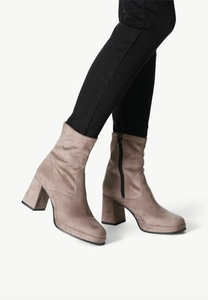 Bottines en suède beige avec un talon carré, une fermeture éclair sur le côté et une texture lisse, associées à un jean skinny noir.