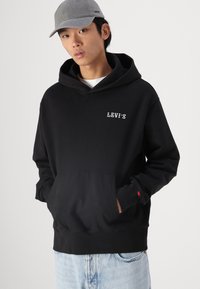 Felpa nera con tasca a marsupio e logo "LEVI'S" bianco. Abbinata con un berretto grigio, dotato di strap regolabile e branding discreto.