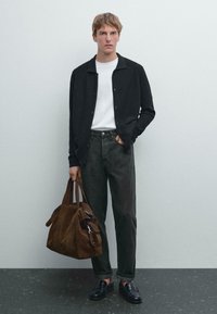 Zwarte button-up cardigan over een wit T-shirt, donkergroene jeans, zwarte instappers en een bruine suède tas met zijaccenten. Gelaagde texturen, casual pasvorm.