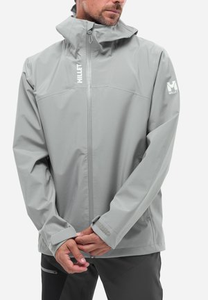 Homme portant une veste imperméable Millet gris clair avec capuche et fermeture éclair devant, ajustant le poignet avec les mains devant.
