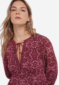 Bordeaux-gepatroneerde jurk met een strikhalslijn en lange mouwen. Bevat een ingewikkeld paisleymotief en subtiele metallic accenten.