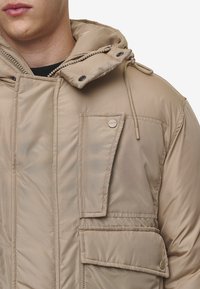 Polstret beige jakke med hette, med glidelås foran, snøring og flere lommer. Jevnt, blankt tekstur med knappdetaljer.