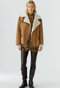 Veste en polaire brune avec un large col, zips argentés, et une robe imprimé léopard en dessous. Le mannequin porte un pantalon en cuir brun ajusté.