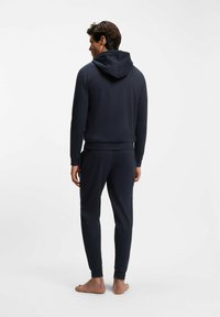 Man som står barfota med ryggen mot kameran, if�örd en mörkblå hoodie och matchande joggingbyxor mot en enfärgad vit bakgrund.