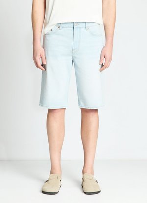 Person steht in hellblauen Jeansshorts, weißem Oberteil und beigen Slip-On-Schuhen vor einfarbig weißem Hintergrund.