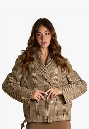 Donna con lunghi capelli castani che indossa un blazer marrone a quadri doppiopetto con cintura e pantaloni marroni abbinati, in piedi su sfondo bianco.