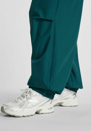 Valkoiset urheilukengät, joissa on verkkokuvioita, päällä vihreänsiniset, elastisilla resoreilla varustetut housut vaaleanharmaalla lattiapinnalla.