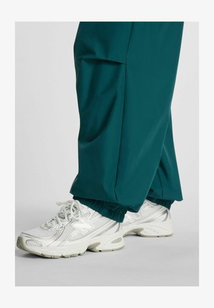 Valkoiset urheilukengät, joissa on verkkokuvioita, päällä vihreänsiniset, elastisilla resoreilla varustetut housut vaaleanharmaalla lattiapinnalla.