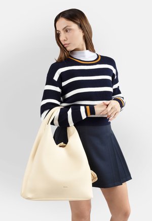 Donna che indossa un maglione e una gonna a righe blu navy e bianche, tiene una grande borsa a tracolla beige e guarda di lato su uno sfondo uniforme.