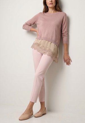 Kvinde, der læner sig mod en væg iført en pink langærmet top med beige blonderkant, lyserøde bukser og beige flade sko.