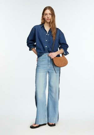 Woman wearing dark blue denim shirt, light blue wide-leg jeans, black sandals, and carrying a brown crossbody bag against a white background.