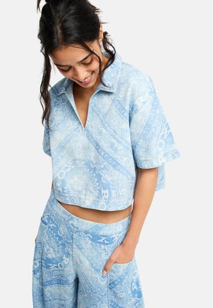 Jeune femme portant une chemise à manches courtes et un pantalon assortis bleu clair à motif, regardant vers le bas et souriant avec une main dans la poche.