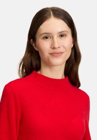 Kobieta z długimi brązowymi włosami, ubrana w jasnoczerwony sweter, z neutralnym wyrazem twarzy i delikatnym makijażem, na jednolitym białym tle.