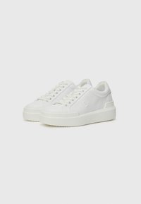 Baskets blanches en cuir lisse avec un bout rond, un laçage à l'avant et une semelle en caoutchouc épaisse. Présence d'un logo discret sur le côté.