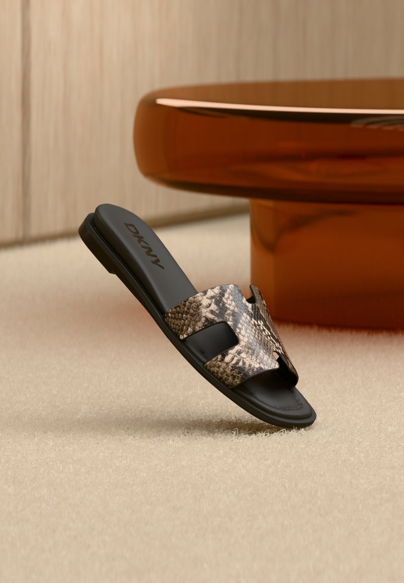 Sandale plate noire avec bride à motif de peau de serpent et logo DKNY, posée sur un tapis beige près d'une table brune brillante.