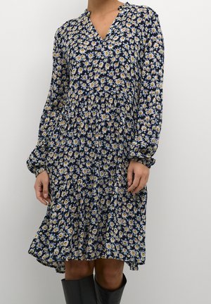 Frau trägt ein langärmliges marineblaues Kleid mit weißem und gelbem Blumenmuster, kombiniert mit schwarzen kniehohen Stiefeln vor einem schlichten Hintergrund.