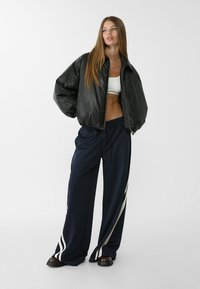 Giubbotto bomber imbottito nero con colletto alto, abbinato a pantaloni felpa blu navy con strisce bianche laterali e vestibilità rilassata. Footwear chunky neri.