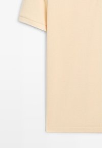 Polo beige à manches courtes en tissu texturé, montrant la partie supérieure gauche et le bord de la manche sur un fond blanc.