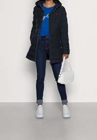 Musta untuvatakki hupulla, sininen t-paita, tummat farkut ja valkoinen tilkkulaukku. Valkoiset lenkkarit täydentävät asun.