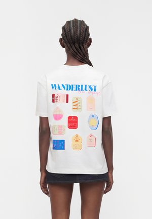 Osoba z warkoczami nosząca białą koszulkę z kolorowymi etykietami podróży i napisem „WANDERLUST Bon Voyage” na plecach.
