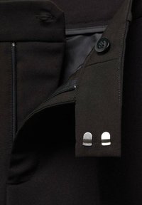 Gros plan d'un vêtement noir avec une fermeture par bouton, deux agrafes argentées à crochet et barre, et un zip partiellement visible.