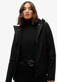 Zwarte softshell jas met een capuchon, voorzien van ritszakken, een gladde textuur en contrasterende rode accenten. Draag het over een zwarte coltrui.