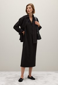 Abito nero al ginocchio con chiusura a bottoni, abbinato a una camicia oversize coordinata. Mocassini neri decorati completano il look.