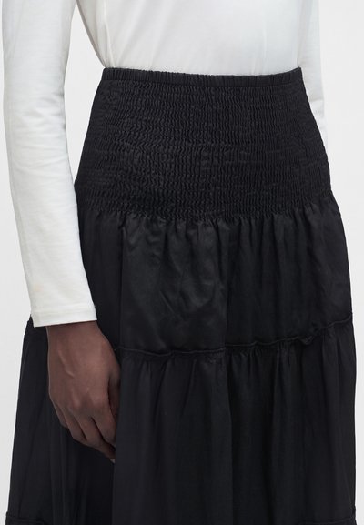 Jupe noire taille haute à volants avec taille élastique froncée, portée avec un haut blanc à manches longues, montrant la taille et la main.