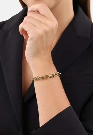Goldene Kettenarmband mit ineinandergreifenden Gliedern, am Handgelenk getragen. Vor einem schwarzen Blazer-Hintergrund platziert, der seinen metallischen Glanz zur Geltung bringt.