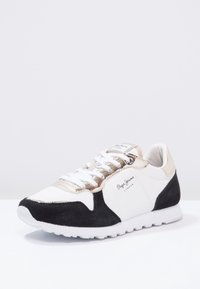 Tenisky s bílou kůží, černými semišovými akcenty a zlatými kovovými detaily. Disponují šněrovacím designem a texturovanou bílou podrážkou.