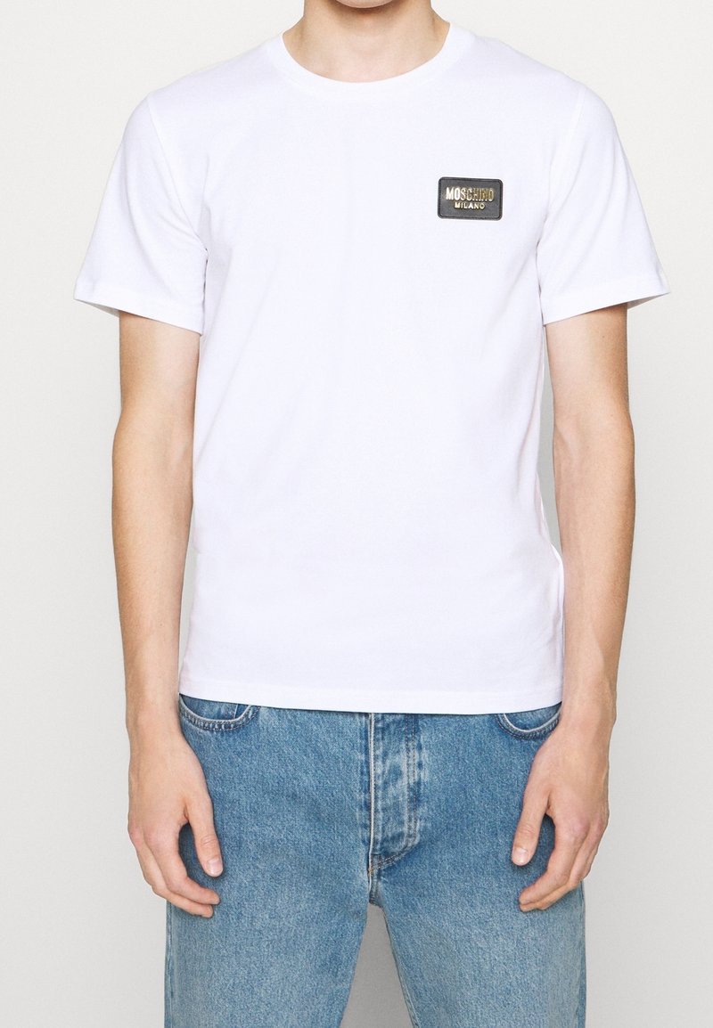 T-shirt en coton blanc à col rond, à manches courtes, avec un patch logo rectangulaire noir sur la poitrine, associé à un jean en denim bleu.