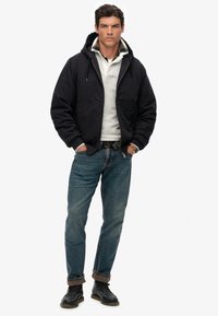 Zwarte gewatteerde jas met een textuurvoering, grijze sweatshirt, blauwe denim jeans en zwarte laarzen, poserend tegen een effen achtergrond.