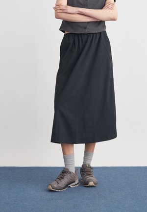 Άτομο όρθιο με σταυρωμένα τα χέρια, φορώντας μαύρη midi φούστα, γκρι αμάνικη μπλούζα, γκρι κάλτσες και γκρι παπούτσια πεζοπορίας σε μπλε πάτωμα.