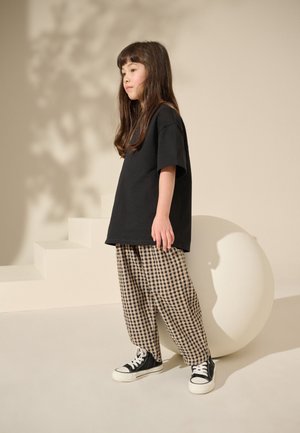 Ein kleines Kind steht seitlich, trägt ein schwarzes übergroßes T-Shirt, karierte weite Hose und schwarze High-Top-Sneakers vor einem neutralen Hintergrund.