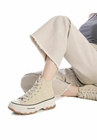 Zapatillas altas de lona beige con suela blanca gruesa, franja negra y puntera redonda. Llevadas con pantalones cortos de color beige claro.