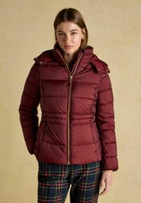 Red puffer jacket with a zip front, flap collar, and gold hardware. Worn over plaid patterned pants. Smooth texture, quilted design.