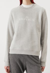 Šedá mikina s kulatým výstřihem, žebrovanými manžetami a lemem, na přední straně s bílým nápisem "HOLZWEILER" přes hrudník, vyrobená z měkké látky.