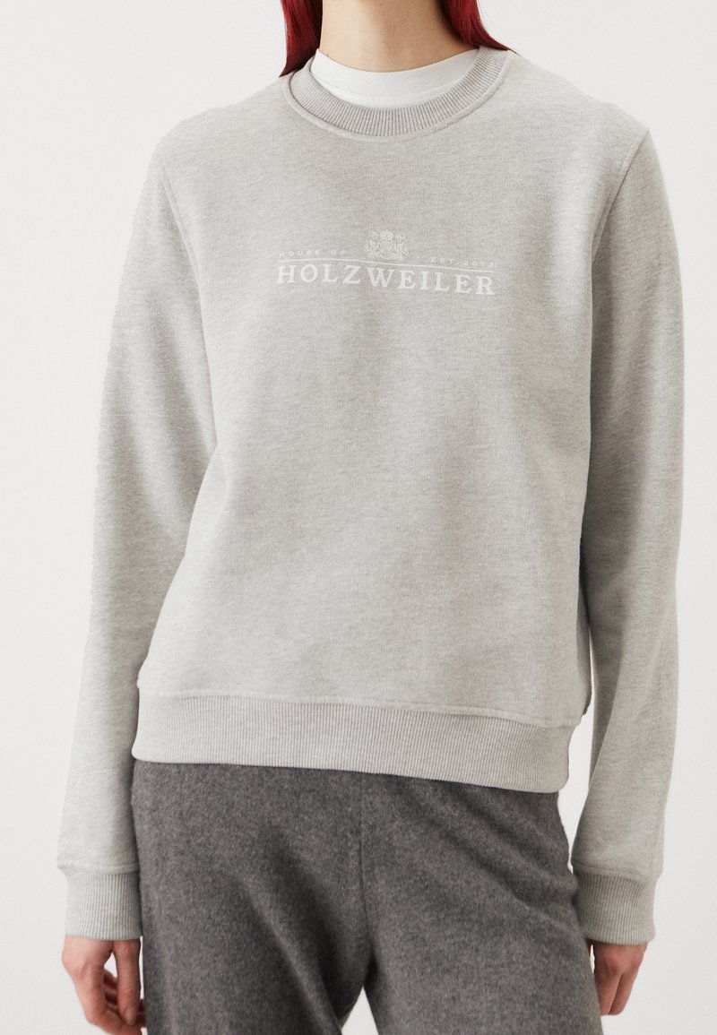 Šedá mikina s kulatým výstřihem, žebrovanými manžetami a lemem, na přední straně s bílým nápisem "HOLZWEILER" přes hrudník, vyrobená z měkké látky.