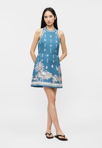 Femeie purtând o rochie albastră fără mâneci cu modele de soare și palmieri, stând desculță pe un fundal alb simplu.