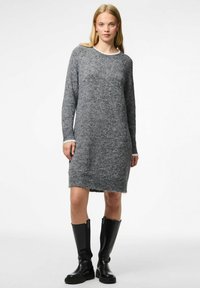 Donna che indossa un lungo abito in maglia grigia abbinato a stivali neri fino al ginocchio, in piedi contro uno sfondo bianco semplice.