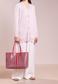 Różowy kardigan w prążki z dekoltem w serek i kieszeniami, zestawiony z czerwoną torbą w wzorki z centralnym paskiem, noszoną na białych szerokich spodniach.