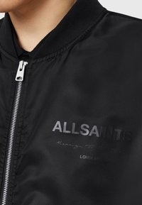 Gros plan sur une personne portant une veste noire avec une fermeture éclair argentée et le texte "ALLSAINTS LONDON" sur la zone gauche de la poitrine.