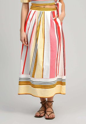 Femme portant une jupe midi à rayures colorées avec des plis, assortie à des sandales plates marron à lanières, debout avec une main dans la poche.