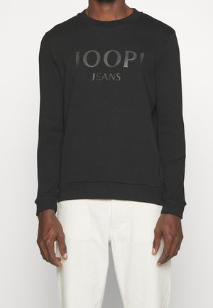 Mężczyzna w czarnym swetrze z długim rękawem z napisem "JOOP! JEANS" i białych spodniach, stojący na jednolitym tle.